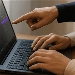 A mentor's hand pointing at a laptop screen while a trainee's hands work the keyboard